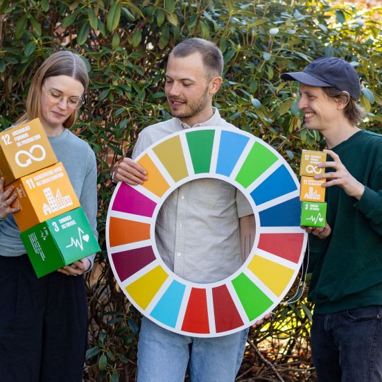 Tre mennesker der står med farvede bokse og FN's Verdensmåls-hjulet